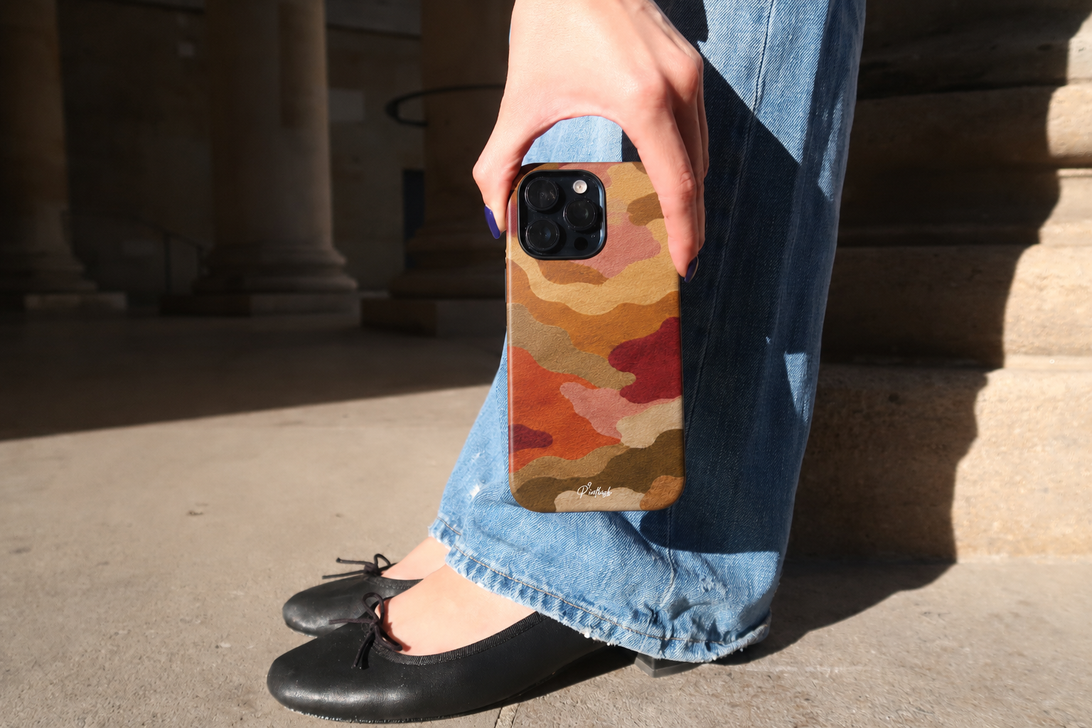 Person holding a phone with a colorful case, wearing jeans and black shoes, outdoors.