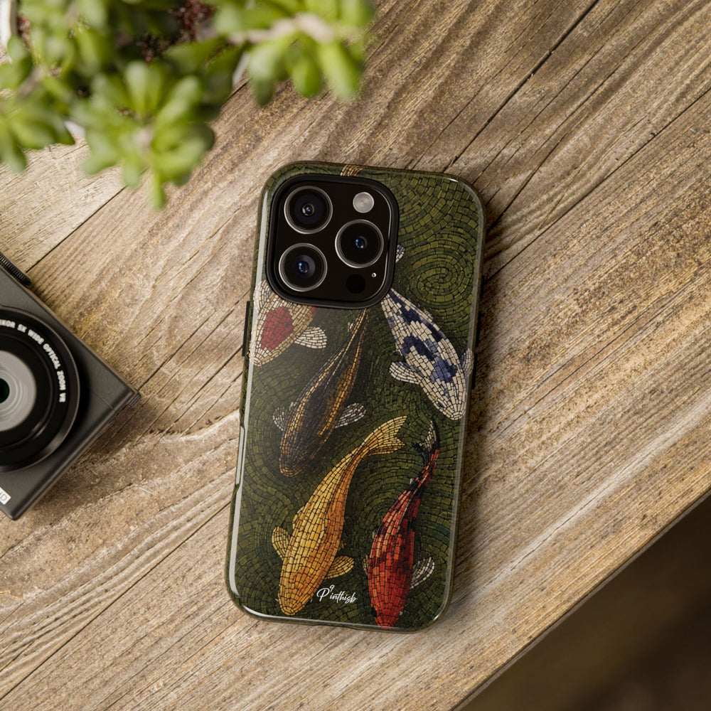 A phone case featuring a vintage-style koi fish illustration in earthy green, gold, red, and blue tones, shown on a wooden tabletop next to a camera and green leaves.