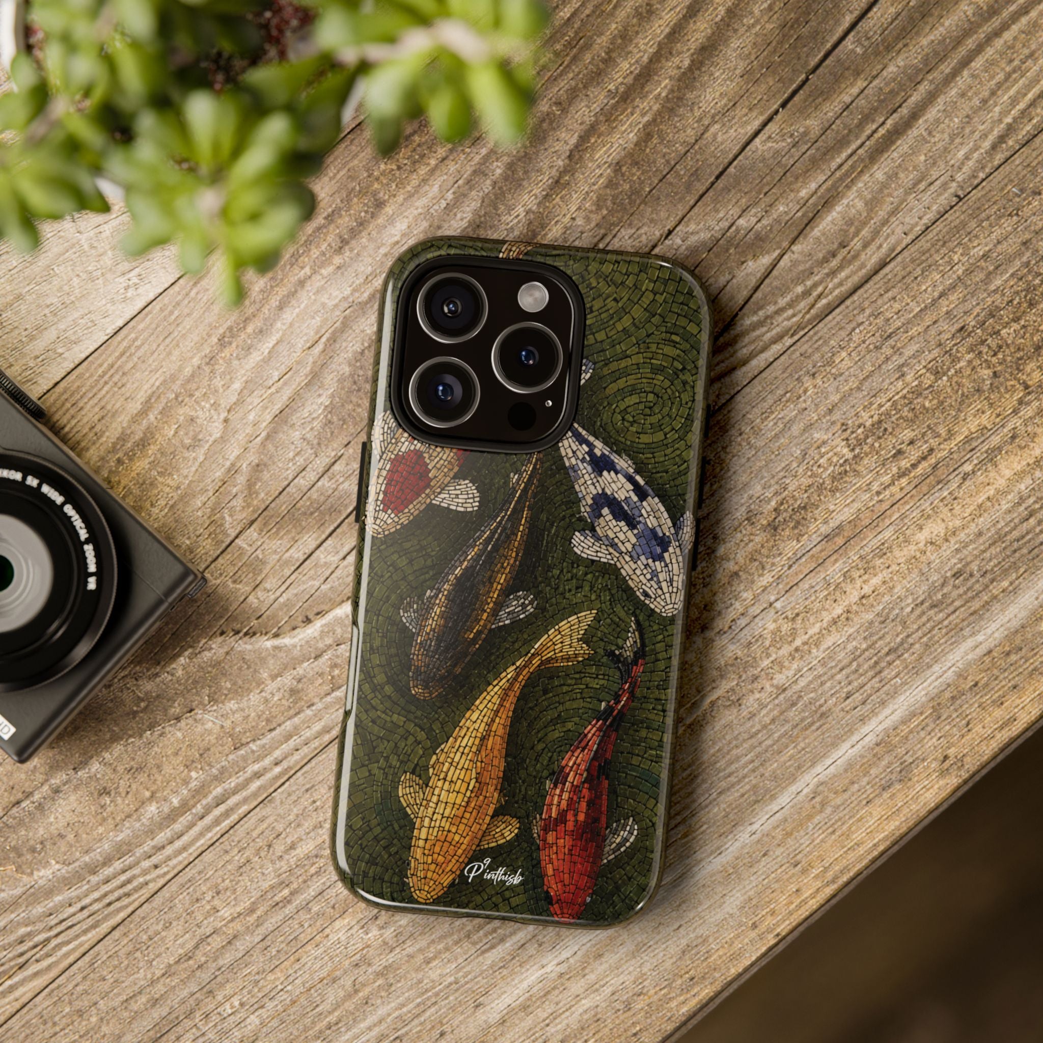 A phone case featuring a vintage-style koi fish illustration in earthy green, gold, red, and blue tones, shown on a wooden tabletop next to a camera and green leaves.