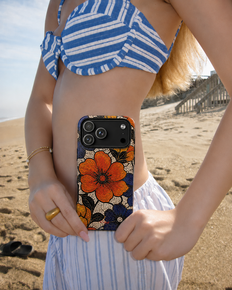 A woman at the beach holding a smartphone with a decorative floral phone case. The case features a large orange flower and blue leaves over a beige mosaic background.