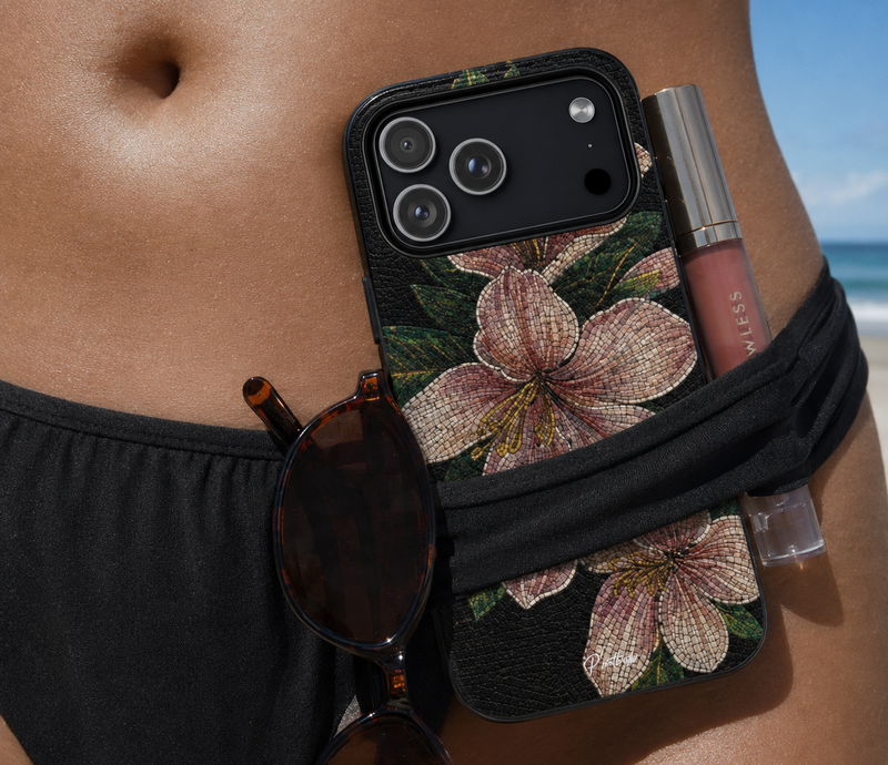 A close-up lifestyle shot of a black phone case with a large pink hibiscus flower print, worn on a model's hip with a black waist bag, sunglasses, and a lip gloss tube.