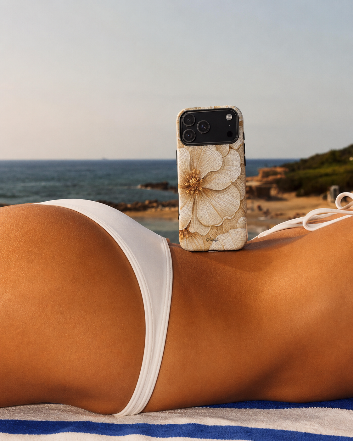 A close-up lifestyle shot of a woman lying on a beach towel by the ocean, wearing a white bikini bottom. A smartphone with a beige floral-patterned case rests on her hip.