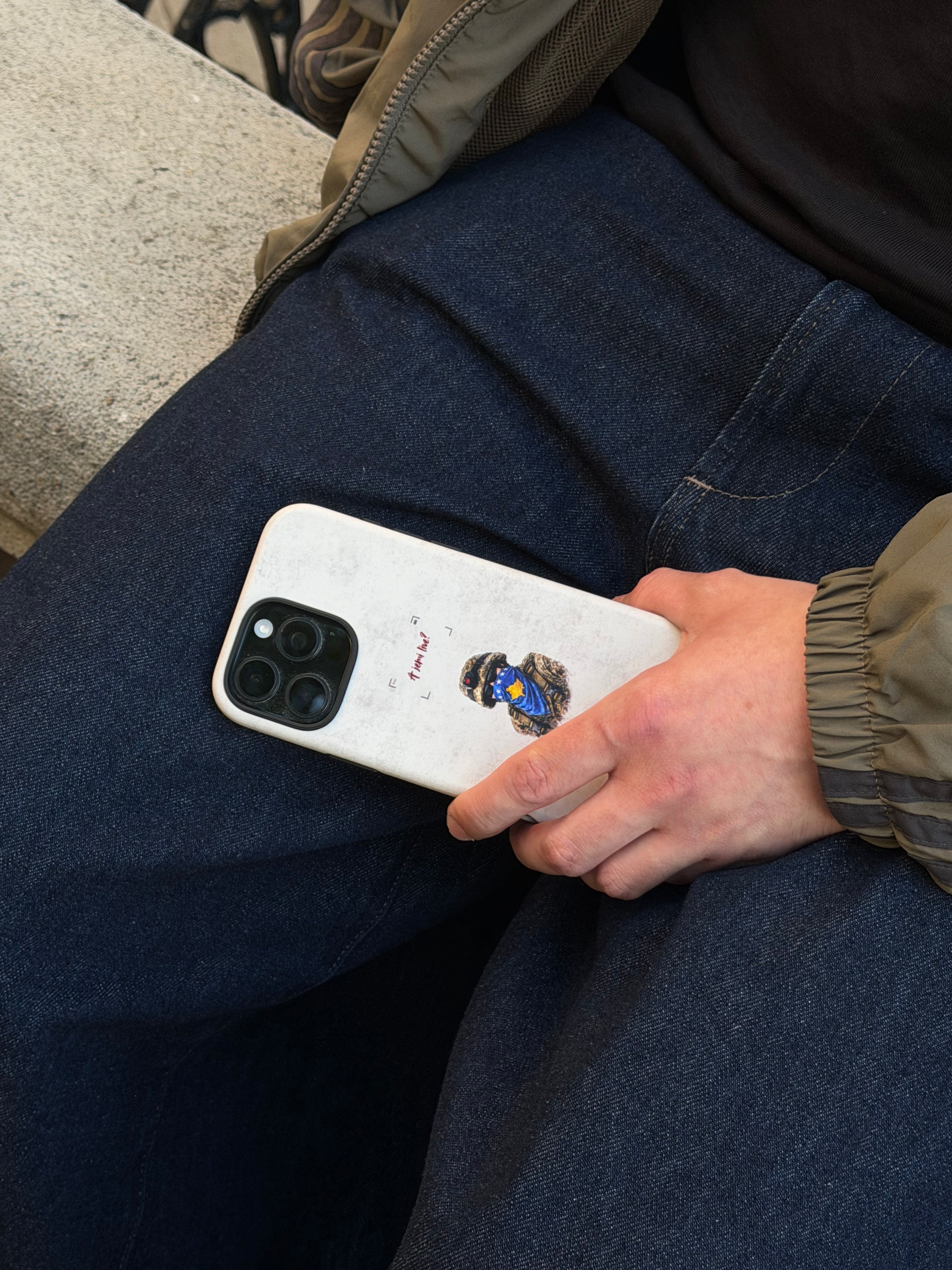 Person holding a white smartphone with a case featuring a kosovan soldier, sitting on a bench.